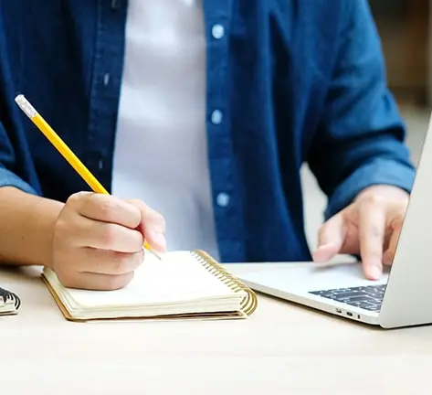 Young university student smiling and diligently working on a laptop with a book, immersed in the research for the scientific thesis in a library. Captures the essence of academic dedication, ideal for those seeking MBA dissertation help and scholarly guidance.